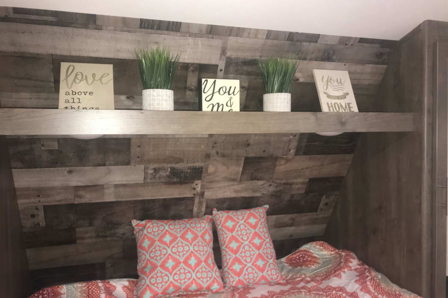 Inside an RV rental: a cozy bedroom with a planked wood headboard, decorative shelf, plants, signs & colorful bedding.