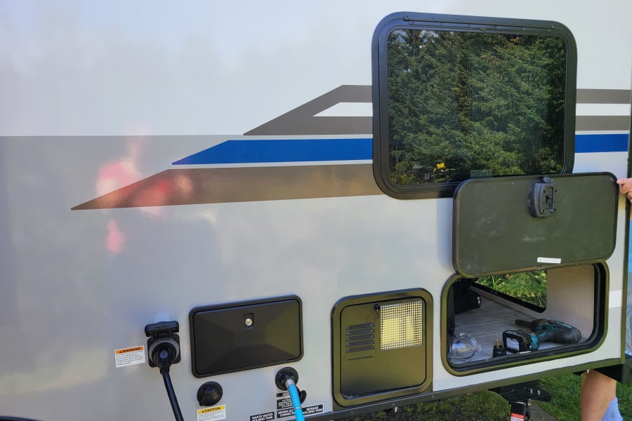 Side of an RV rental showing utility hookups (power, water, sewer) and an open storage compartment with tools, ready for camping.