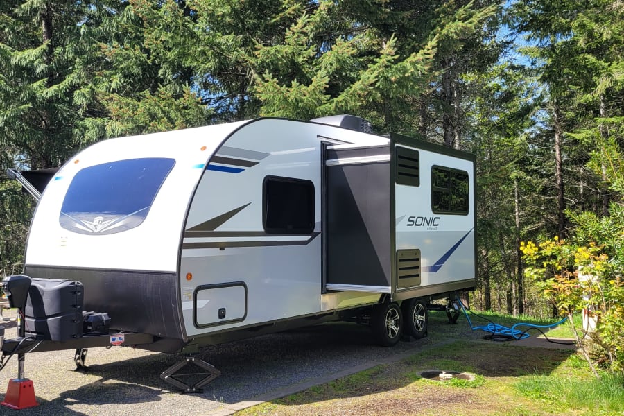 This rv rental, a white Sonic travel trailer, is set up at a wooded campsite on a sunny day. Connections visible.