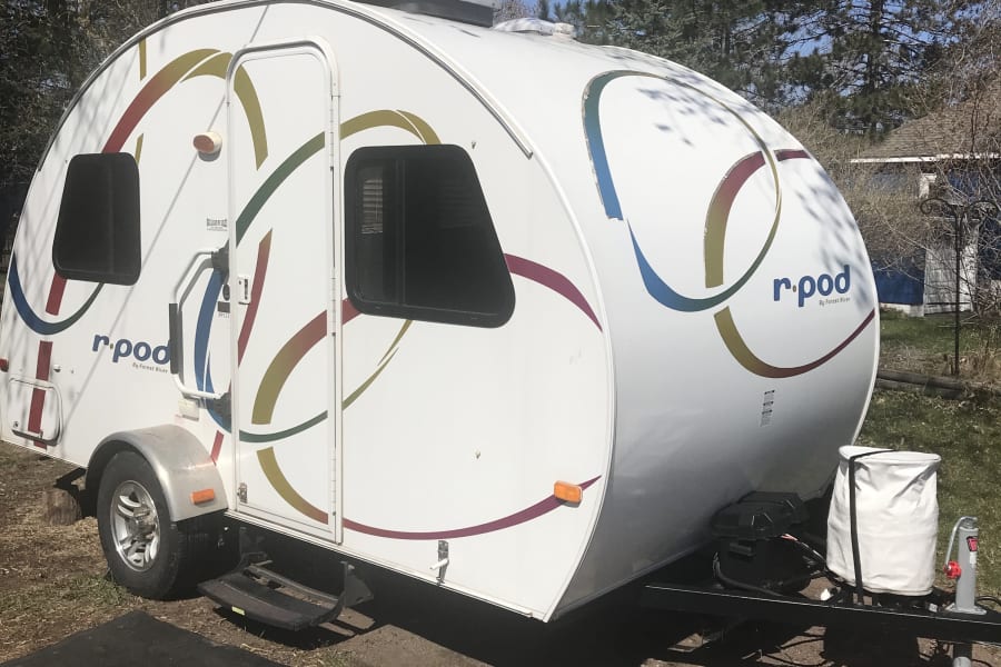 White r-pod teardrop RV rental, decorated with colorful swirls, parked outdoors on gravel. Perfect for camping adventures.