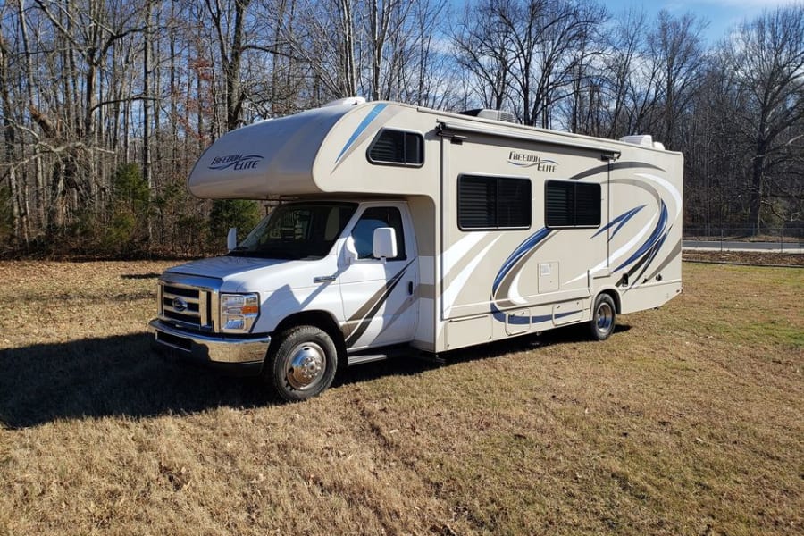 This beige and white Freedom Elite Class C motorhome, ideal for an rv rental, is parked outdoors on dry grass.
