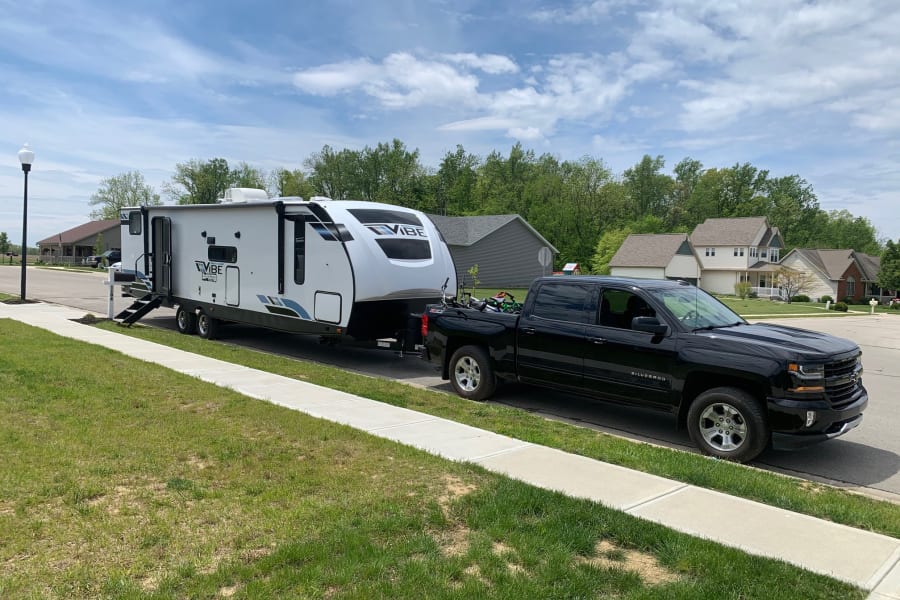 Black pickup truck towing a white VIBE travel trailer, an RV rental, parked on a suburban street.