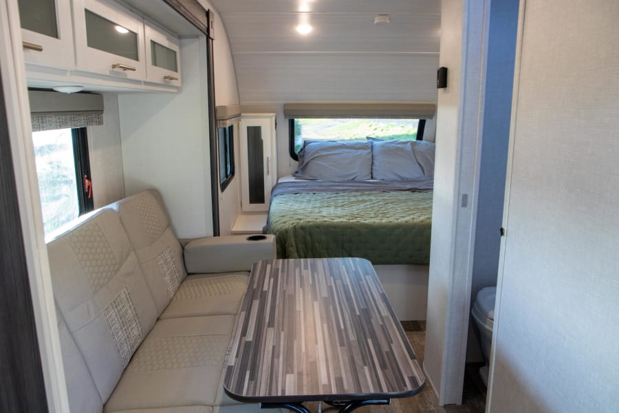 RV rental interior featuring a light-colored sofa, dining table, bed with green comforter, and bathroom access.