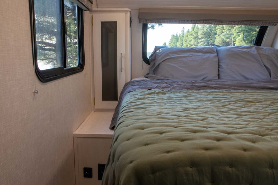 Cozy RV rental bedroom with a green quilted bed, gray pillows, bedside cabinet, and large windows showcasing outdoor trees.