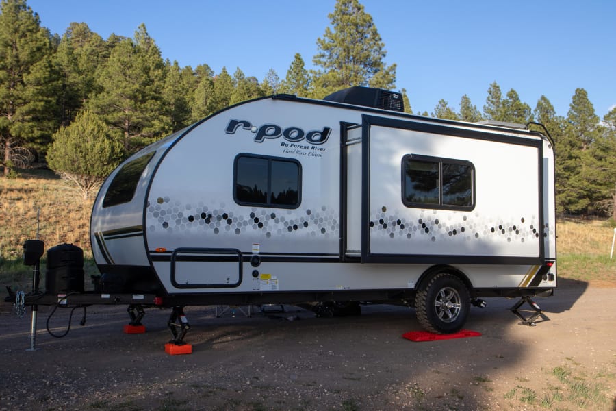 White R-Pod travel trailer, great for an rv rental, parked and leveled at a sunny campsite near pine trees.