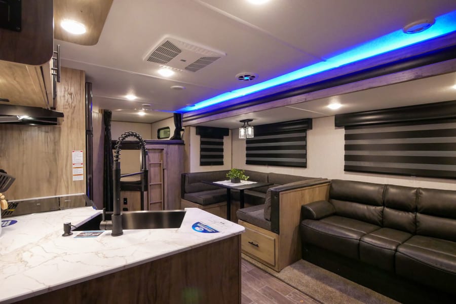 Modern RV rental interior: kitchen, cozy dinette, sofa, and bunks under blue lights.