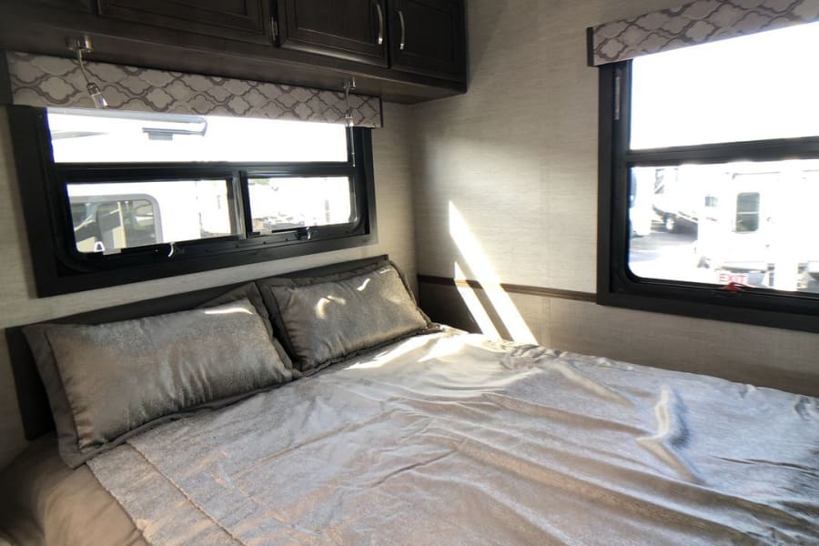 Spacious RV rental bedroom with a comfortable bed, multiple windows, and dark wood overhead cabinets. Bright and inviting.