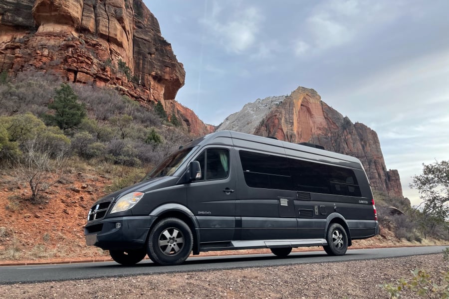 A sleek black RV rental van on a scenic road, flanked by majestic red rock formations under an overcast sky.