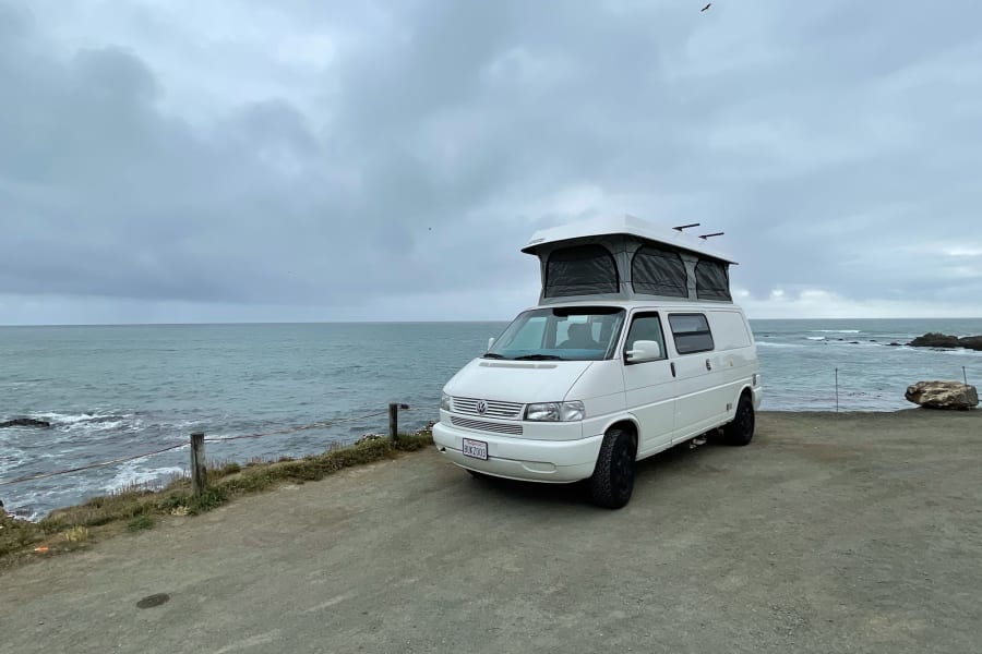 White VW pop-top camper van RV rental, parked oceanfront under a cloudy sky.