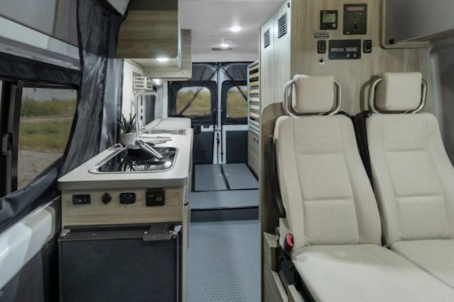 Modern RV rental interior featuring a kitchenette with sink, stove, and fridge, plus comfortable light-colored passenger seats.