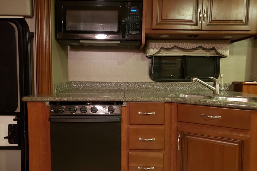 RV rental kitchen: black microwave, stove/oven, stainless sink, ample wood cabinets, and speckled counter.