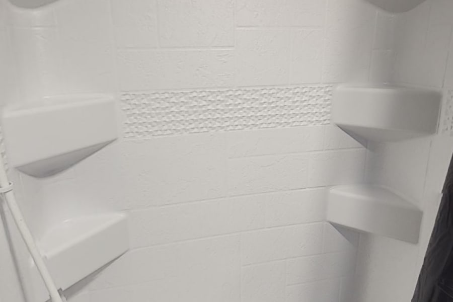 Clean white RV rental shower with tub, built-in shelves, and handheld sprayer. Compact for travel needs.
