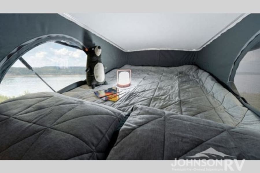 Experience the comfort of an RV rental: a cozy bedroom with quilted bedding, lamp, plush toy, and a beautiful waterside view.