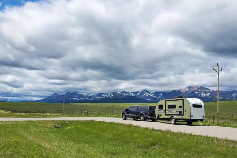 Scenic **rv rental** & truck parked by a field, with snow-capped mountains under a cloudy sky. Ideal for a mountain road trip.