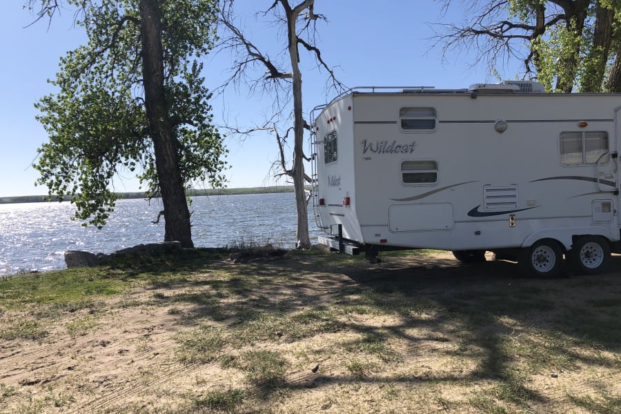 White Wildcat rv rental parked on sandy ground beside a sunlit lake and trees, perfect for a peaceful camp.