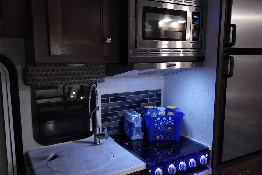 RV rental kitchen: A compact galley with stainless microwave, stove (blue lights on knobs), sink, and fridge. Fully equipped.