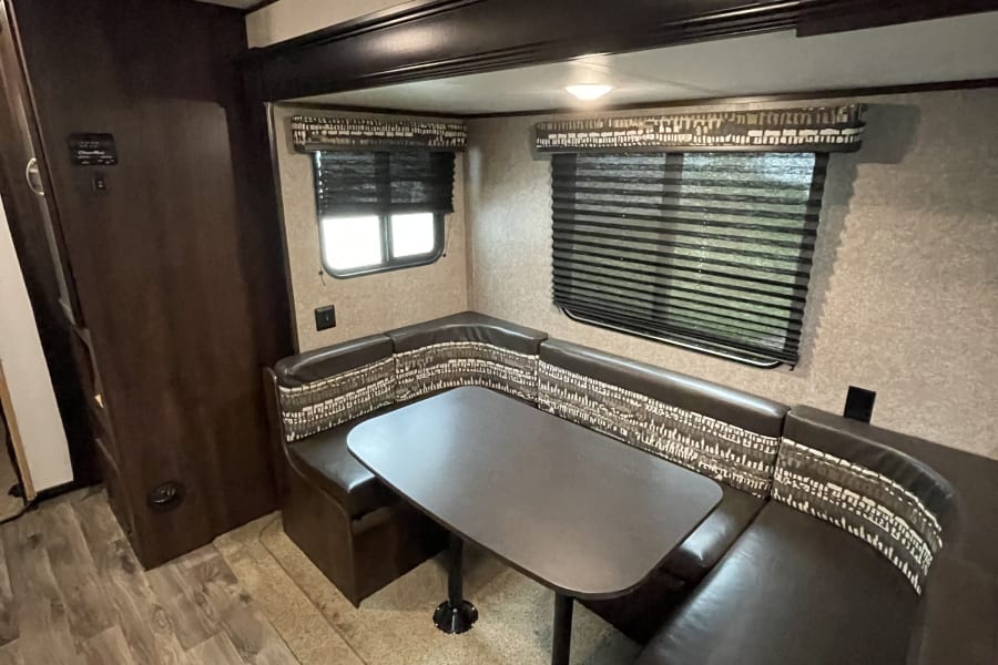 An RV rental's cozy dinette with U-shaped brown seating, a large table, and window blinds, ready for your next adventure.