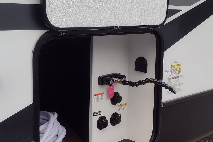 Open utility hatch on an RV rental, revealing water connections, a spray hose, and an electrical cord.