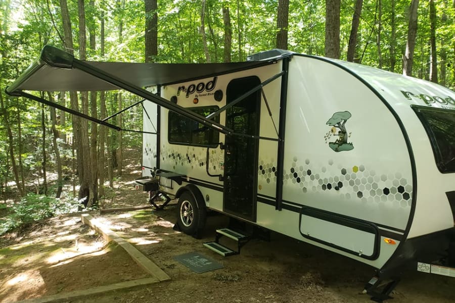 White r-pod RV rental under a deployed awning in a shady forest campsite. Features honeycomb patterns & tree decal.