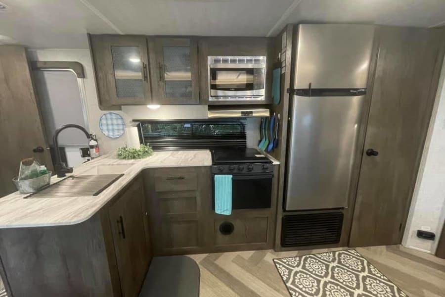 A compact RV rental kitchen featuring a stainless steel fridge, microwave, stove, sink, and wood-look flooring.