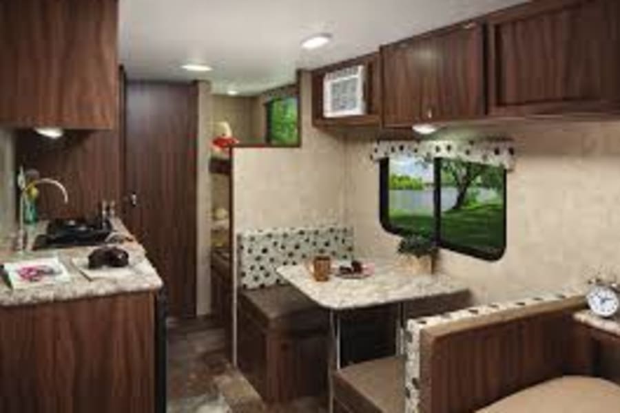 This RV rental interior shows a compact kitchen, dinette, and bunk beds. Scenic lake views from windows, dark wood cabinets.