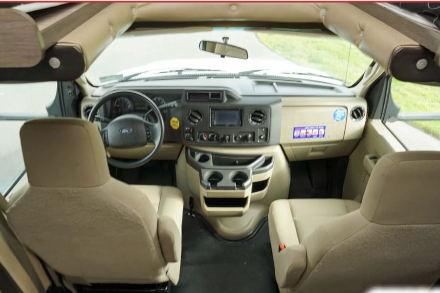 View of an RV rental's front cabin. It shows the driver's cockpit, dashboard with a Ford steering wheel, and two tan seats.