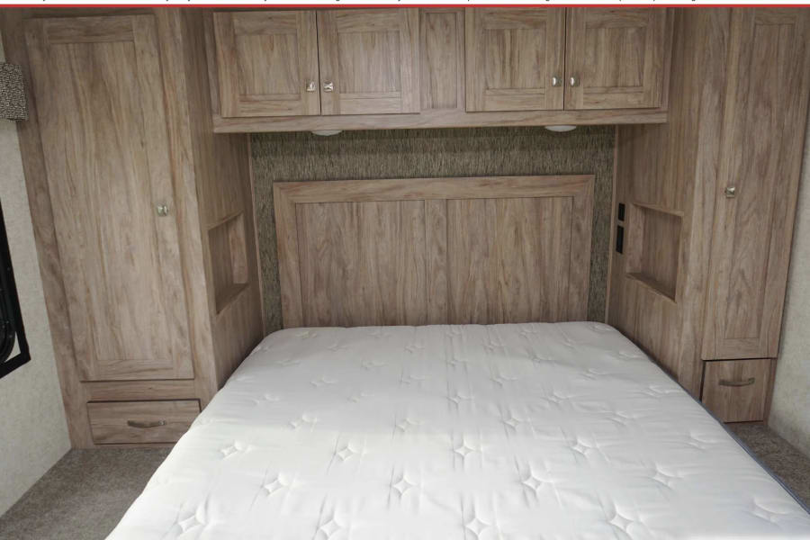 RV rental bedroom: white mattress, wooden headboard, overhead cabinets, bedside storage nooks & wardrobes, plus windows.