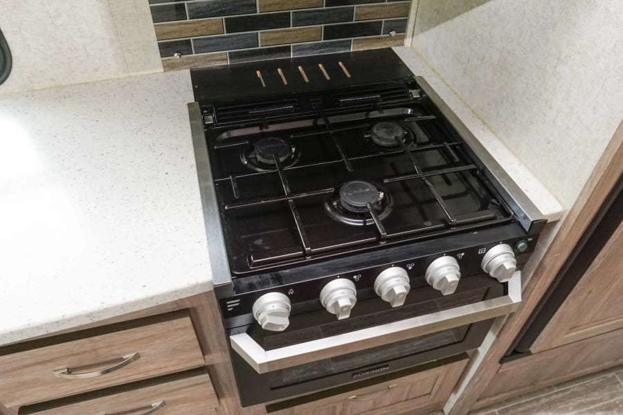 This RV rental features a modern kitchen with a shiny stainless steel sink, a black gas stove/oven, and light wood cabinets.