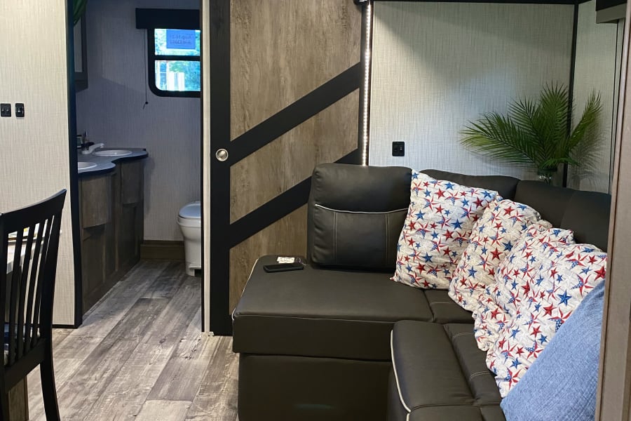 Modern RV rental interior with a dark sectional sofa and star pillows. A sliding barn door opens to the bathroom. Wood floors.