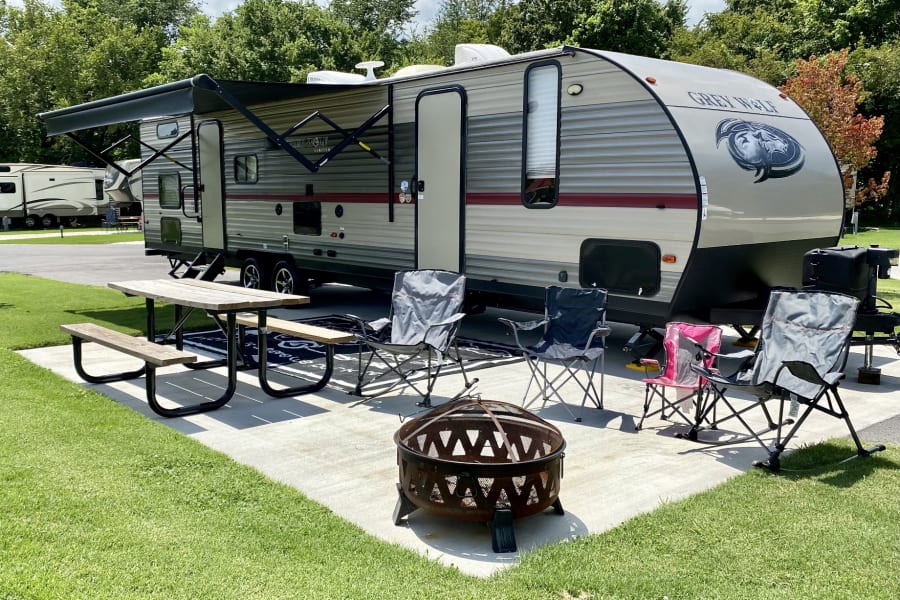 A Grey Wolf rv rental travel trailer, with awning out, is parked at a sunny campground site, beside a picnic table, fire pit, and chairs on a patio.