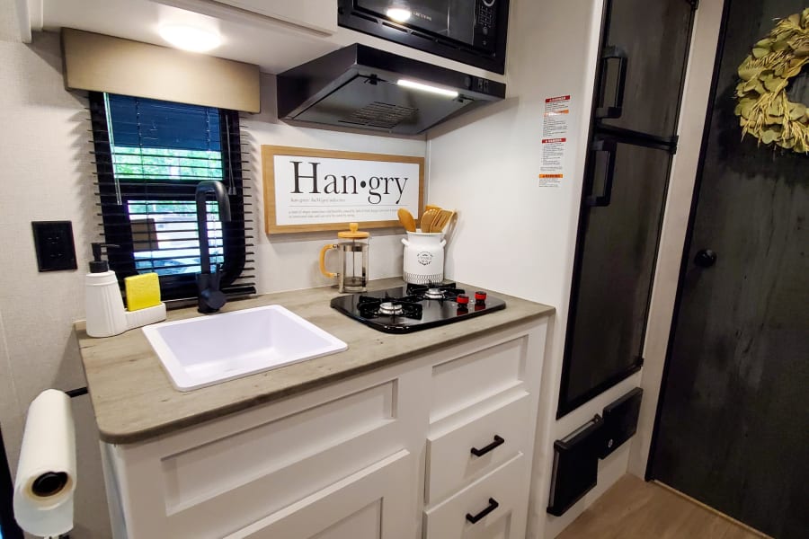 This RV rental features a compact, modern kitchen with white cabinets, sink, stove, microwave, and fridge, ideal for on-the-go meals.