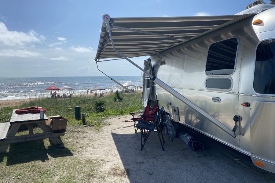 Coastal rv rental: Silver Airstream camper under a striped awning, parked beachfront. People and umbrellas on the sand.