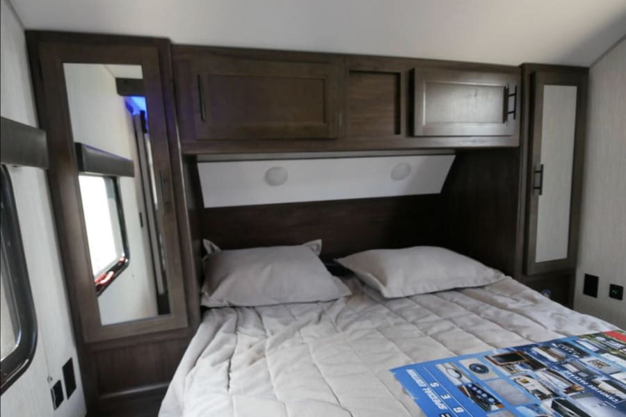Cozy RV rental bedroom featuring a compact bed with pillows, overhead cabinets, and mirrored wardrobes for a functional space.