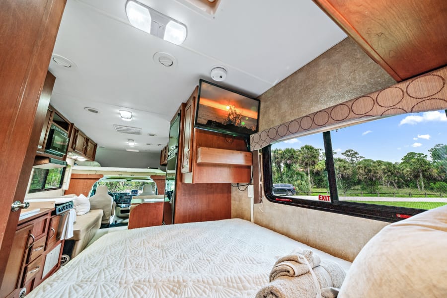 Comfortable rv rental interior with a cozy bed, mounted TV, kitchen, and scenic window view, ideal for travel.