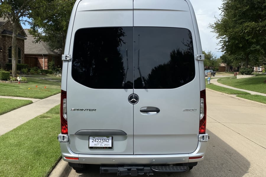 Rear view of a silver Mercedes Sprinter van RV rental parked on a tree-lined suburban street with green lawns.