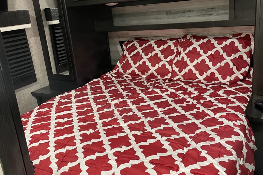 Inside a comfortable RV rental bedroom, featuring a vibrant red and white patterned bed, dark wood cabinets, and shelving.