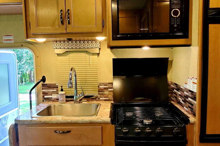 RV rental kitchen: light wood cabinets, stainless sink, black stove & microwave. Includes AC & roof vent for comfort.