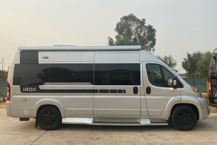 A silver IROK camper van, ideal for your next rv rental adventure, is parked on concrete under a hazy sky.