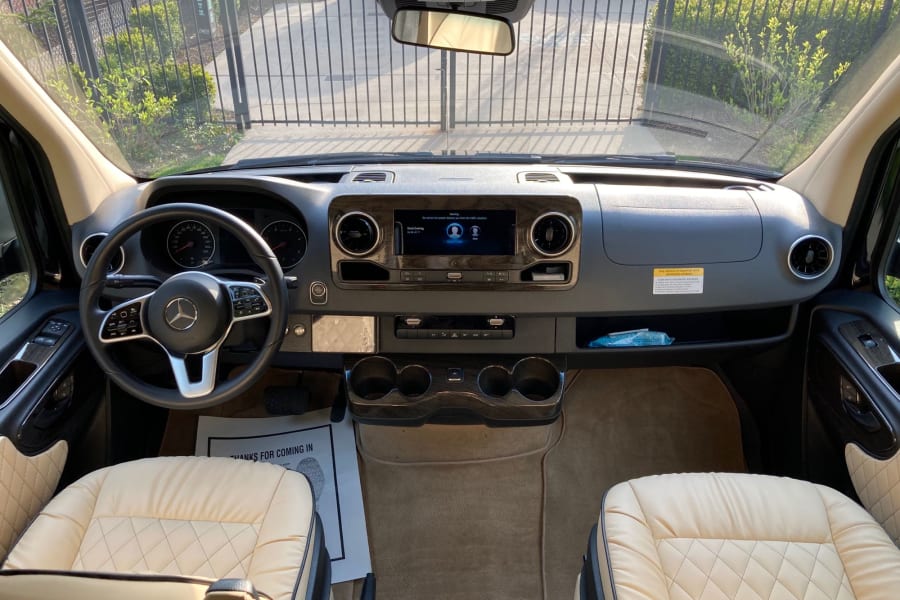 Luxury RV rental's detailed interior. View of the front cabin with Mercedes dashboard, quilted leather seats, and gate outside.