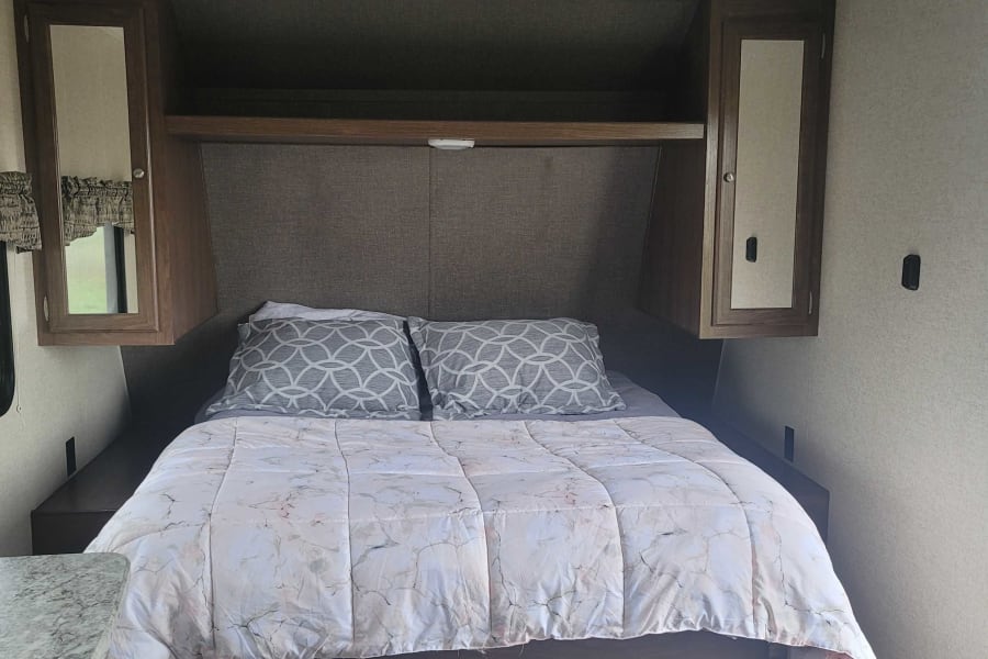 A tidy RV rental bedroom with a bed, light comforter, gray pillows, and dark wood cabinetry. Features overhead storage and ceiling lights.