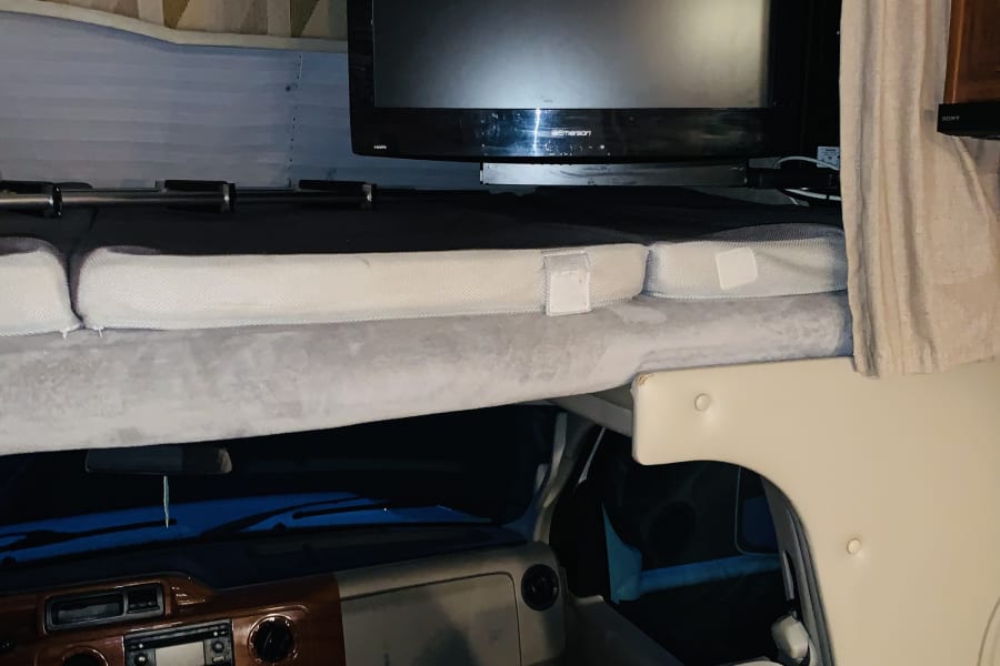 Inside an RV rental: the view of the overhead bed with a TV mounted above, and the vehicle's dashboard below.