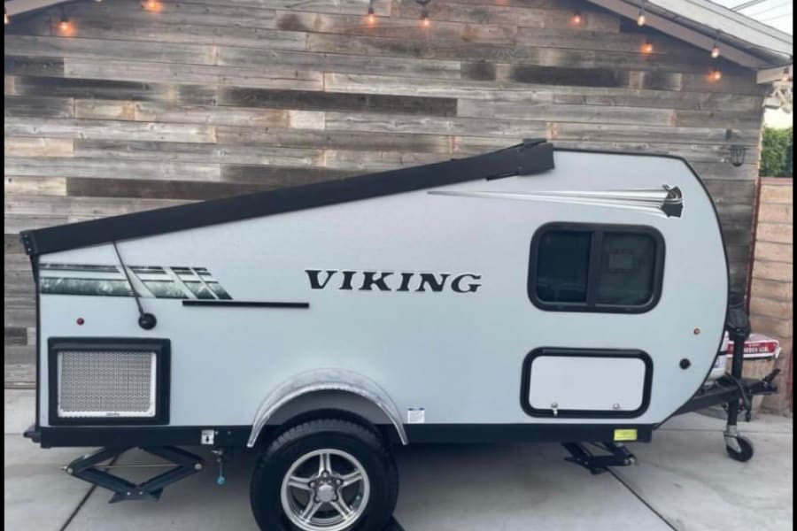 This light gray Viking teardrop RV rental awaits adventure, parked against a wooden wall with festive lights.