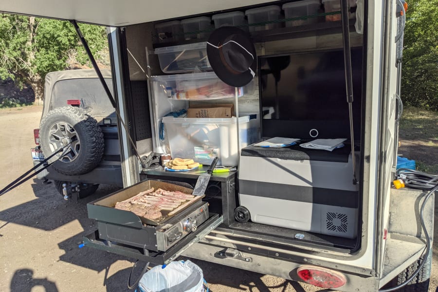 RV rental outdoor kitchen with a Blackstone griddle cooking bacon, storage, and portable fridge, perfect for camping meals.