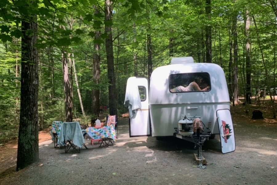 A person relaxes inside a white teardrop RV rental parked in a shaded forest campsite with a picnic area.