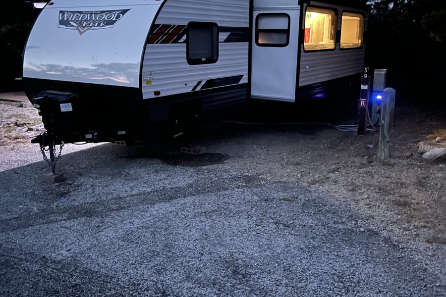 Wildwood X-Lite rv rental travel trailer parked at a campsite at night, hooked up to power with interior lights on.