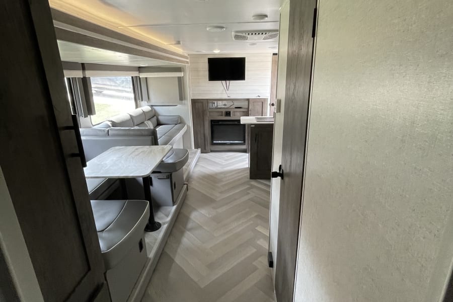 Bright RV rental interior shows a hallway leading to a modern living area with couch, dinette, and fireplace.