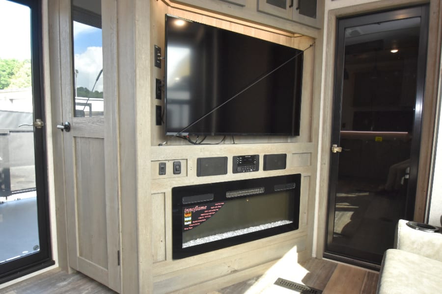 RV rental entertainment center with a large TV, fireplace, and integrated audio system, set in light wood paneling.