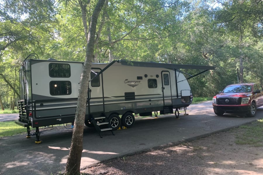 This gray and white RV rental, with its red tow truck, is fully set up at a sunny, wooded campsite, awning extended.