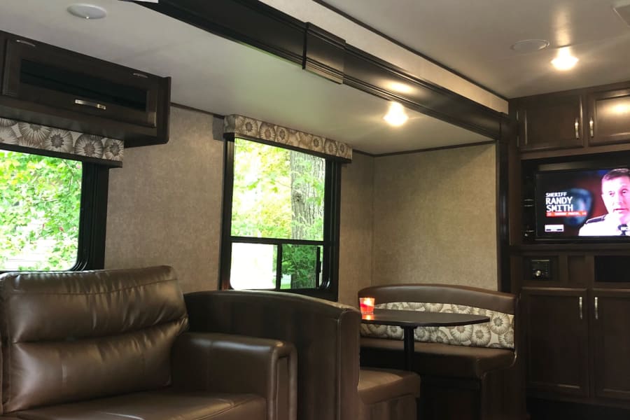 Cozy RV rental interior with a brown sofa, dinette, TV, and "this is us" family photo decor. Ideal for trips.