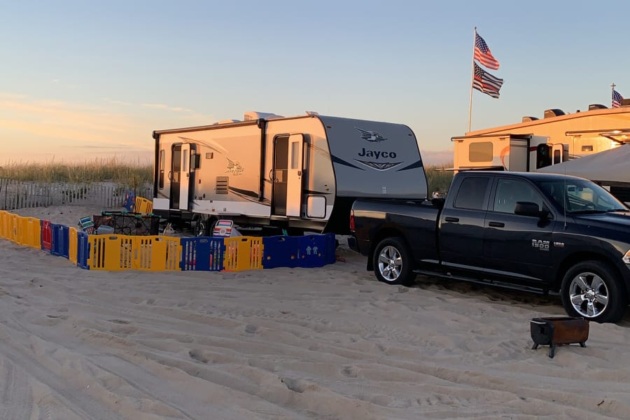 An RV rental and truck are set up for beach camping on a sandy shore at sunset. Flags fly near other campers.